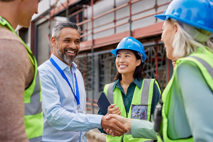 Smiling construction professionals exchanging a handshake as part of their partnership on site. Happy engineer shaking hands at construction site with happy businessman. Handshake between construction manager with architect at building site, conclude an agreement.